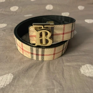 Burberry vändbart bälte med guldspänne - Snyggt vändbart bälte från Burberry med klassiskt rutigt mönster i beige, rött och svart på ena sidan och svart präglat skinn på andra. Stort guldigt B-spänne framtill. Perfekt accessoar för att lyfta din outfit med lyxig känsla.