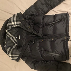 Svart pufferjacka/väst från Burberry - Svart pufferjacka från Burberry med klassiskt rutigt foder i grått och vitt. Jackan har dragkedja, knappar och flera fickor med dragkedja. Materialet är polyester och jackan har en skön, vadderad känsla som håller dig varm. Perfekt för kalla dagar och snygg streetstil.