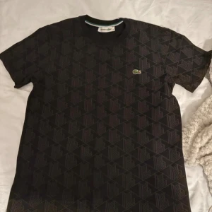 Svart mönstrad t-shirt från Lacoste monogram - Snygg svart t-shirt från Lacoste med geometriskt mönster över hela plagget och klassisk krokodillogga på bröstet. T-shirten har rund hals och korta ärmar, perfekt för dig som gillar stilrena detaljer och ikoniska märken.