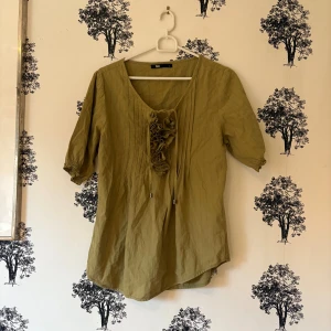Olivgrön blus med volang från Flash - Olivgrön blus från Flash i storlek S med korta ärmar och v-ringad hals. Framtill finns en snygg volangdetalj och tunna dekorativa veck. Blusen har avslappnad passform och är gjord i ett lätt bomullsmaterial, perfekt för vår och sommar.