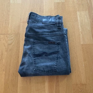 Mörkgrå jeans från Nudie Jeans - Snygga mörkgrå jeans från Nudie Jeans med klassisk femficksdesign och svarta detaljer på bakfickan. Jeansen har rak passform och är tillverkade i mjukt denimtyg. Perfekt för dig som gillar stilrena och tidlösa jeans.