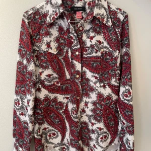 Paisleymönstrad skjorta Isabel Marant - Paisleymönstrad skjorta från Isabel Marant i vitt med röda och grå detaljer. Skjortan har klassisk krage, långa ärmar och knappar framtill samt på manschetterna. Snygga bröstfickor med lock och en loose passform. Perfekt för dig som gillar statement-plagg.