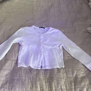 Vit ribbad blus från Zara - Säljer en vit ribbad blus från Zara i storlek M. Toppen har långärmade ärmar, rund halsringning och knappar framtill. Materialet är mjukt och stretchigt, perfekt för en chill look. Croppad passform som ger en snygg siluett.