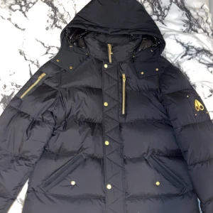 Svart dunjacka Moose Knuckles - Svart dunjacka från Moose Knuckles med guldfärgade detaljer och dragkedjor. Jackan har huva, flera fickor med knappar och en snygg logga på ärmen. Perfekt för kalla dagar med sin tjocka och quiltade design. Insidan har glansigt foder och tydlig Moose Knuckles-märkning.Helst i Norrköping kontakta.Den är XL men passar L