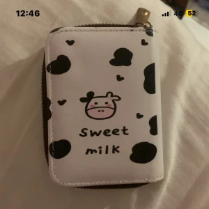 Söt plånbok med ko-motiv - Plånbok i vitt med svarta fläckar och gulligt ko-ansikte på framsidan. Texten 'sweet milk' och små hjärtan ger en lekfull vibe. Stängs med dragkedja och har en kompakt, smidig form som passar perfekt i fickan eller väskan.