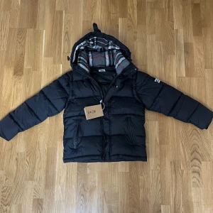 Svart pufferjacka från Burberry - Svart pufferjacka från Burberry med huva och rutig innerfoder i grått, vitt och rött. Jackan har två sidofickor med dragkedja och en tydlig Burberry London England patch på ärmen. Perfekt för kalla dagar och har en stilren, modern look.