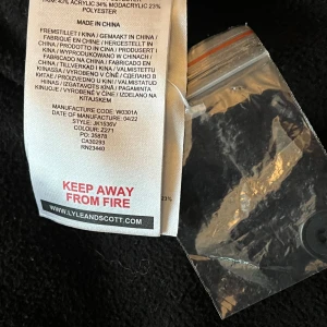 Svart parkajacka med fuskpäls från Lyle & Scott - Parkajacka från Lyle & Scott i svart med huva och beige päls. Jackan har dragkedja och knappar framtill, samt Lyle & Scotts klassiska gula logga på bröstet. Materialet är polyester och akryl, perfekt för kalla dagar. Nypris 2400