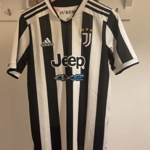 Juventus hemmatröja Adidas 15-16 år/ S - Juventus officiella hemmatröja från Adidas med klassiska svarta och vita ränder, broderat klubbmärke med tre stjärnor och Jeep-sponsor på bröstet. Tillverkad i lätt och ventilerande Primegreen-material. Perfekt för dig som älskar fotboll och vill visa ditt stöd för Juve.
