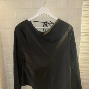 Svart omlottopp från Gina Tricot - Snygg svart omlottopp från Gina Tricot i glansigt satinliknande material. Toppen har lång ärm, djup draperad rygg med snörning och en cool omlott-look framtill. Perfekt för dig som gillar stilrena och trendiga plagg med lite extra edge.