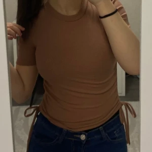 Ribbad nude topp med snörning från Zara - Säljer en ribbad nude topp från Zara med korta ärmar och rund hals. Toppen har snygga snörningsdetaljer på båda sidor som går att justera för en mer figurnära passform. Perfekt att styla med jeans eller kjol för en chill look.