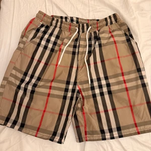 Burberry mönstrade bad byxor  - Säljer ett par snygga shorts med klassiskt rutigt mönster i beige, svart, vitt och rött. De har elastisk midja med vit snörning och sidofickor. Perfekta för chill dagar och har en riktigt clean streetstil.
