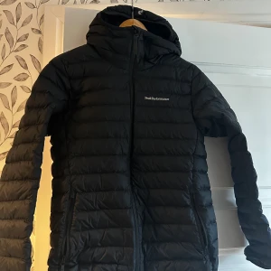 Svart dunjacka Peak Performance S - Svart dunjacka från Peak Performance i storlek S. Jackan har huva, två sidofickor med dragkedja och quiltad design. Perfekt för kalla dagar och har en stilren look med diskret logga på bröstet och bak på huvan. Inga anmälningar på varken tyg eller dragkedja!