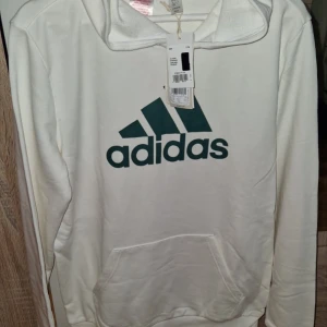 Vit hoodie från Adidas med grön logga - Snygg vit hoodie från Adidas med stor grön logga på bröstet. Tröjan har huva, magficka och långa ärmar. Perfekt för dig som gillar sportig och clean stil. Materialet är mjuk bomull som känns skönt mot huden.