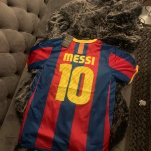 Barcelona Messi 10 fotbollströja  - Barcelona hemmatröja med Messi och nummer 10 på ryggen. Klassisk design med breda blå och röda ränder, gula detaljer och klubbmärke på bröstet. Tillverkad i lätt polyester med Dri-FIT-teknologi för skön känsla på planen. Perfekt för dig som älskar fotboll och Messi.
