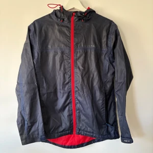 Mörkblå vindjacka från Everest - Snygg mörkblå vindjacka från Everest med röd dragkedja och detaljer. Jackan har huva med snörning, lång ärm och är tillverkad i lätt polyester. Perfekt för blåsiga dagar och har en sportig vibe med kontrastfärgad insida.