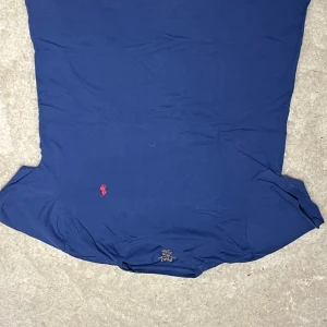 Blå t-shirt från Polo Ralph Lauren - Snygg blå t-shirt från Polo Ralph Lauren med klassisk röd broderad logga på bröstet. Modellen har rund hals och korta ärmar, tillverkad i mjuk bomull som känns skön mot huden. Perfekt för dig som gillar stilrena och ikoniska plagg.