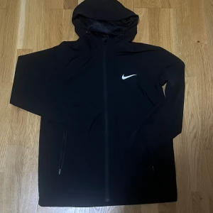 Svart vindjacka från Nike med huva - Svart vindjacka från Nike med huva och dragkedja framtill och är emot vatten . Den passar xs/s helt ny utan prislapp den passa inte mig.