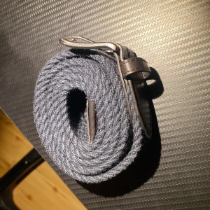 Blått stickat bälte med skinnspänne - Snyggt blått stickat bälte med mörkbruna skinn-detaljer och metallspänne. Bältet har en flätad struktur som ger en cool och modern look. Perfekt för att lyfta din outfit med en unik accessoar. Passar dig som gillar att sticka ut med stilrena detaljer.