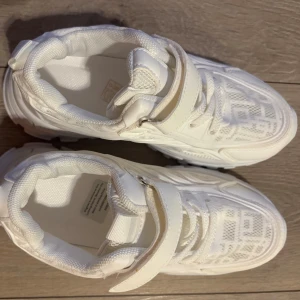 Helt nya oanvända Vita chunky sneakers med kardborre stl 34 - Säljer ett par vita chunky sneakers med både snörning och bred kardborreband över foten. Skorna har en ovandel i mesh och syntet för extra ventilation. Rund tå och platt sula ger en sportig vibe. Perfekt för dig som gillar stilrena och bekväma sneakers. Den är märkt som stl 36 men den är en 34