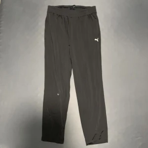 Svarta träningsbyxor från Puma - Svarta träningsbyxor från Puma med elastisk midja och diskret logga på vänster lår. Byxorna har en rak passform och är gjorda i ett lätt syntetmaterial som andas bra. Perfekta för gymmet eller sportaktiviteter.