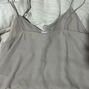 Beige linnetopp med volang Gina Tricot - Supersöt beige linnetopp från Gina Tricot med tunna axelband och en snygg volangdetalj längs ringningen. Toppen har en enkel och stilren design som är lätt att matcha med olika outfits. Perfekt för dig som gillar en clean och feminin look. Varan har inga tecken på användning 