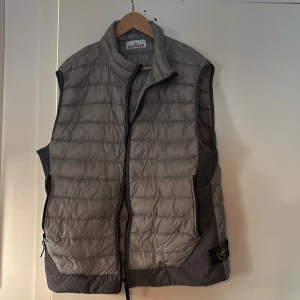 Grå dunväst från Stone Island - Snygg grå dunväst från Stone Island med quiltad design och hög krage. Västen har dragkedja framtill, två sidofickor med dragkedja och den klassiska Stone Island-loggan på sidan. Perfekt lager-på-lager-plagg med lätt och smidig känsla.