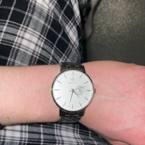Silverfärgad klocka från Gant - Stilren klocka från Gant med rund, vit urtavla och minimalistisk design. Klockan har en silverfärgad länk i metall och diskret datumvisning. Perfekt accessoar för dig som gillar klassisk och clean stil. Enda nackdelen är att klockvisarna är lösa ( billigt att fixa kanske 150kr). Klockan har ett nytt batteri. Den är i mycket bra skick annars. Pris går att diskuteras. Har även bitar, så ifall klockan är för liten kan du göra den större :)