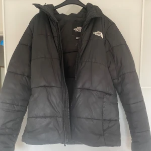 Svart pufferjacka från The North Face - Säljer en svart pufferjacka från The North Face i storlek M. Jackan har en klassisk quiltad design, huva och dragkedja framtill. Perfekt för kalla dagar och riktigt snygg streetstil med logga på bröstet.
