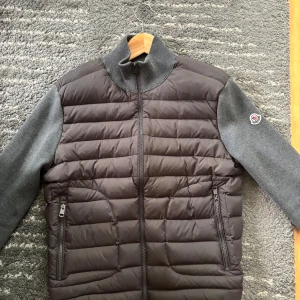 Grå Moncler Maglione Tricot Cardigan - Snygg pufferjacka från Moncler i mörkgrått med quiltad front och stickade ärmar och rygg. Jackan har hög krage, dragkedja framtill och två fickor med dragkedja. Klassisk Moncler-logga på ena ärmen. Perfekt för dig som vill ha en stilren och exklusiv look.