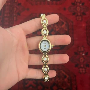 Guldig armbandsklocka med pärldetaljer - Snygg armbandsklocka från Howdy med guldig metall och vita pärldetaljer i länkarna. Oval urtavla med pärlemor och små glittriga stenar som index. Klassisk och elegant design som sticker ut på handleden. Klockjusterar följer med!