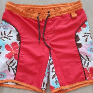 Badshorts - Säljer ett par färgglada badshorts från Body Zone i storlek 128. Shortsen har röd och orange bas med blommigt mönster i rosa, vitt, brunt och blått på sidorna. De har snörning i midjan och är perfekta för strand eller pool. Materialet är lätt och snabbtorkande. Har en fläck.