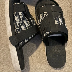 Svarta Dior sandaler med mönster - Säljer ett par svarta sandaler från Dior med ikoniskt mönster i vitt och svart. Sandalerna har breda remmar med snörning och en platt sula. Materialet är en mix av textil och syntet, med Dior-logga på sulan och remmen. Perfekt för dig som vill ha en lyxig vibe.