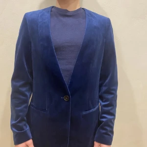 Blå sammetskavaj med enkel knäppning - Snygg kavaj i djupblå sammet med en knapp framtill och två fickor. Klassisk modell med smal krage och lång ärm, perfekt för dig som vill sticka ut med en lyxig känsla. Passar till både skjorta och t-shirt.