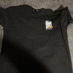 Svart Casa Blanca t-shirt med tryck - Svart t-shirt från Casa Blanca med färgglatt tryck fram och stort motiv på ryggen. Trycket visar en tropisk scen med palmer och texten 'Casa Blanca'. Klassisk rund hals och korta ärmar. Perfekt för dig som gillar streetwear med unik design.