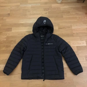 Svart dunjacka från Sail Racing - Svart dunjacka från Sail Racing med huva och dragkedja | Använd ett fåtal gånger men har en liten reva på ryggen (se bild) | Storlek 160 | Nypris 2000 mitt pris 479