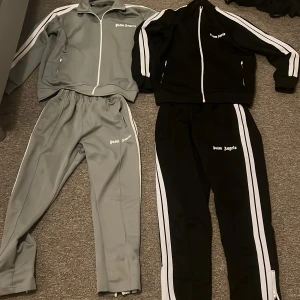 Palm Angels träningsbyxor. Gröna setet i storlek L) och det svarta i storlek M - Säljer två par Palm Angels träningsbyxor, ett par i grått och ett par i svart, båda med vita sidoremsor och logga på låret. Byxorna har elastisk midja, raka ben och dragkedja vid benslut. Perfekta för en sportig och trendig look. ”Gröna settet är i storlek L” och den svarta ”M” men kan sitta som s  också. Kom med bud