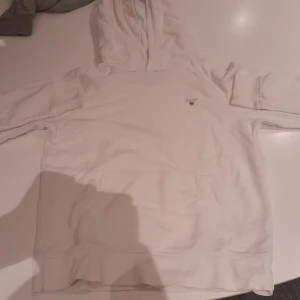 Vit hoodie från GANT med huva - Vit hoodie från GANT med klassisk huva och magficka. Tröjan har diskret GANT-logga på bröstet och är tillverkad i mjuk bomull. Perfekt för en chill och stilren look.