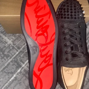 Christian Louboutin svarta sneakers  - Svarta sneakers från Christian Louboutin med coola svarta nitar på tån och klassisk röd sula med logga. Skorna har snörning och är gjorda i mocka med rund tå. Medföljer dustbag och extra nitar. Perfekt för dig som vill sticka ut med lyxig streetstyle.