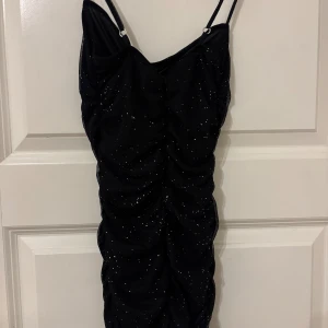 Svart glittrig kort klänning Bershka - Supersnygg svart kort klänning från Bershka med tunna axelband och glittriga detaljer. Klänningen har rynkade partier framtill som ger en cool och trendig look. Perfekt för nyår