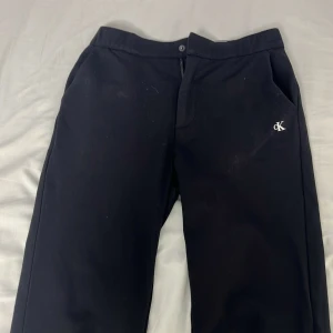 Svarta byxor från Calvin Klein Jeans - Svarta byxor från Calvin Klein Jeans med diskret CK-logga broderad vid fickan. Klassisk rak passform och midja med knapp och dragkedja. Snygga och stilrena, perfekta för dig som gillar enkel design och vill ha ett par byxor som funkar till allt.