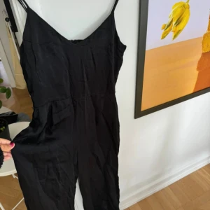 Svart byxdress  - Svart byxdress från H&M med smala axelband och v-ringning. Byxdressen har en enkel och stilren design, perfekt för dig som gillar minimalistiska plagg. Tillverkad i ett mjukt tyg som faller snyggt längs kroppen.