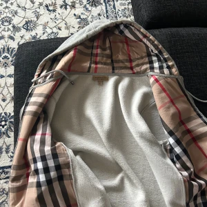 Grå teddyjacka från Burberry med rutig huva - Säljer en grå teddyjacka från Burberry med ikonisk rutig huva i beige, svart och rött. Jackan har dragkedja, snörning vid huvan och mjukt fleecefoder. Perfekt för dig som gillar klassisk stil med en twist. Burberry-logga broderad på bröstet.