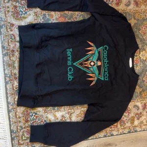 Svart sweatshirt Casablanca Tennis Club - Svart sweatshirt från Casablanca med färgglatt tryck på bröstet där det står 'Tennis Club' och 'Casablanca' i grönt och orange. Tröjan har rund hals, långa ärmar och är gjord i mjuk bomull. Perfekt för dig som gillar streetwear med sportig vibe.