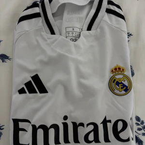 Real Madrid vit matchtröja Adidas S - Snygg Real Madrid fotbollströja i vit med svarta detaljer och klubbmärke på bröstet. Adidas-logga och sponsortryck på framsidan, samt texten '¡Hala Madrid!' i nacken. Kortärmad modell i lätt och ventilerande polyester, perfekt för träning eller match.