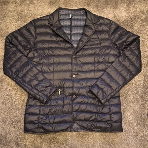 Mörkblå dunjacka från Strömberg - Snygg svart dunjacka från Strömberg i kavajmodell med knappar framtill och klassisk krage. Perfekt för dig som vill ha en stilren look med extra värme. Kontakta vid frågade.