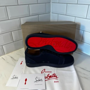 Mörkblå sneakers från Christian Louboutin - Snygga mörkblå sneakers från Christian Louboutin med ikonisk röd sula och broderad logga på hälen. Skorna har rund tå, snörning och är tillverkade i ett lyxigt skinnmaterial. Perfekt för dig som vill sticka ut med exklusiv stil och detaljer.