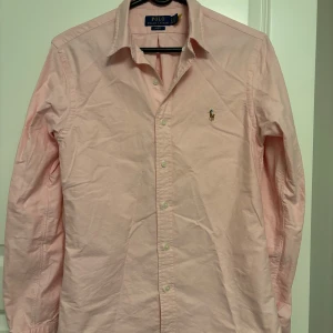 Rosa skjorta från Polo Ralph Lauren - Säljer en stilren rosa skjorta från Polo Ralph Lauren i slim fit. Skjortan har klassisk krage, långa ärmar och knäppning framtill. Broderad logga på bröstet och tillverkad i mjuk bomull. Perfekt för dig som gillar en clean och snygg look.