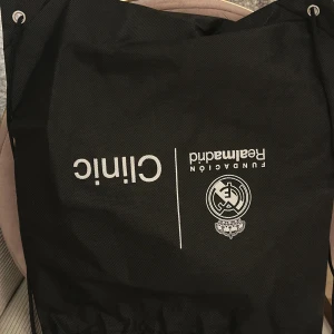 Svart Real Madrid gympåse Clinic - Svart gympåse med Real Madrid Fundación Clinic-tryck i vitt. Väskan har snörning upptill och är tillverkad i slitstark syntet. Perfekt för att bära träningskläder eller skor. Enkel och stilren design med klubbens logga framtill.