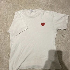Vit Comme des Garçons Play t-shirt - Vit t-shirt från Comme des Garçons Play med det ikoniska röda hjärtat med ögon broderat på bröstet. Klassisk passform och rund halsringning, tillverkad i mjuk bomull. Perfekt för en clean och stilren look.