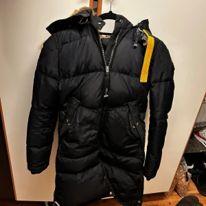 Mörkblå Parajumpers dunjacka med päls - Säljer en mörkblå lång dunjacka från Parajumpers med fluffig päls runt huvan och gul detaljrem vid axeln. Jackan har dragkedja, knappar och två stora fickor framtill. Perfekt för kalla vinterdagar och riktigt snygg med sin mörka färg och coola patch på ärmen. Kan behövas en snabb tvätt. Skick syns på bilderna. Qr kod och taggarna till den finns kvar som visas på bilden 